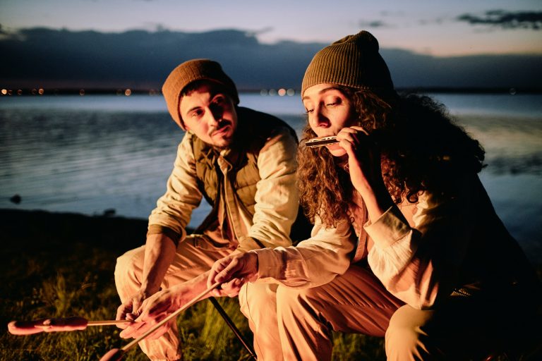 Young male hiker looking at his wife playing harmonica in front of campfire